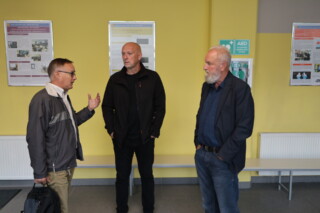 Trije moški stojijo na hodniku v polkrogu. Eden moški govori, druga dva ga poslušata.