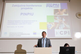Moški v sivi obleki in s temnimi lasmi stoji za govrniškim odrom in se nasmiha. Za njim je veliki pano s predstavitvijo na kateri je napis Zaključna konferenca projekta PINPIU.