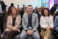 Tri osebe sedijo v prvi vrsti konferenčne dvorane, pred njimi so priponke z imeni in gradiva. V ozadju so drugi udeleženci in snemalna oprema.