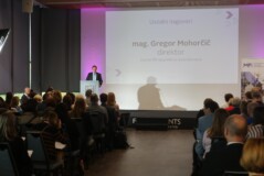 Govorec stoji na odru pred polno dvorano poslušalcev. Na projekcijskem zaslonu je napis »Uvodni nagovori« in ime govorca, mag. Gregor Mohorčil, nazivom direktor Centra RS za poklicno izobraževanje.