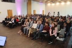 Širok pogled na konferenčno dvorano, kjer je več deset udeležencev sedečih v vrstah. V ospredju so zasloni z imenom govorca, v ozadju pa snemalna ekipa n kamere.