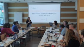 Predavateljica predstavlja vlogo raziskovalnega inštituta na področju izobraževanja. Udeleženci spremljajo predstavitev v konferenčni dvorani.