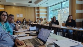 Udeleženci sedijo za konferenčnimi mizami in pozorno spremljajo predstavitev. V ospredju so odprti prenosni računalniki in delovni materiali.